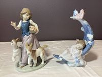 Two Lladro porcelain figurines placed side by side, one of a child asleep on crescent moon and the other girl with two dogs