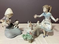 Two Lladro porcelain figurines side by side on a surface against a purple wall. Left figurine shows a girl holding a cat with a dog sitting nearby. Right figurine depicts a girl giving milk to cats beside a curved bench.