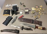 Full overview of all items on table including knives, tools, flask, binoculars, pocket watch, pins, buttons, and patches