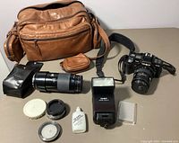 Full lot contents including brown leather carrying case, Minolta camera with lens mounted, zoom lens, flash unit, lens cleaning fluid, and lens caps spread on a table.