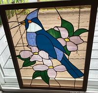 Full view of stained glass window showing blue jay and pink flowers with green leaves, framed in wood and hanging by chains.