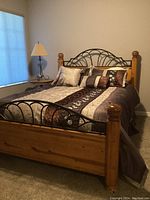 Queen bed fully assembled showing wooden posts, iron headboard and footboard, with bedding and pillows, beside window with lamp on side table visible.