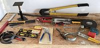 Photo showing all tools laid out on plywood surface including stapler, cutters, tape measures, pliers, measuring square, metal stand, strap wrench