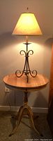 Photo of vintage wooden round side table with lamp on top, showing iron base lamp with cream shade and wooden table with pedestal and small drawer, natural wood color with no finish.