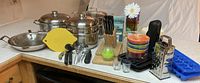 Photo of assorted kitchen cookware and utensils on counter including pots, pans, knives, grater, cutting boards, bowls, thermos, ice cube trays, and measuring cups.