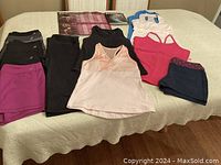 Photo showing several pieces of women's athletic clothing including multiple sleeveless tops in pink, peach, white with blue back, and black, along with shorts and pants in black, navy, and magenta colors laid out on a bed.