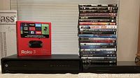 Photo shows Roku 3 player in its original box, Motorola PVR DCX3400-M cable box, Dynex DVD player, and a stack of 20 DVDs in their cases.