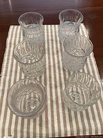 Top view and side angle of six clear cut glass juice or water glasses arranged on a striped cloth. One glass shows a small crack near the rim.