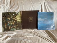 3 LP record covers laid out on fabric surface, Creedence Clearwater Revival album with motion blur cover, Crosby Stills Nash & Young brown/black album, and Eagles blue Hotel California cover.