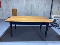 Full view of the harvest table showing natural pine wood top with black painted legs against a gray floor and unfinished wall background.