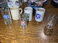 Six beer mugs displayed on wooden surface, featuring 3 stoneware mugs and 3 glass mugs with various brewery logos