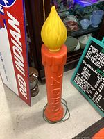 Front view of red-orange candle with yellow flame on top, showing NOEL lettering and electric cord at base.