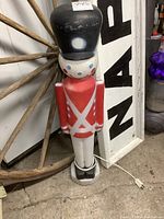 Full front view of 30 inch vintage blow mold nutcracker soldier showing red coat, white pants, tall black hat with white and blue markings, on a white and black base.