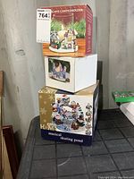 Stacked boxes of three Christmas-themed candle holders including a musical skating pond and Santa sleigh scene.