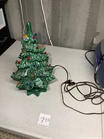 Ceramic green Christmas tree with multi-colored bulb decorations, power cord and plug visible on table.