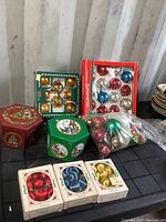 Overview of six boxes and loose bags of Christmas tree balls in various colors and packaging styles on a black surface.