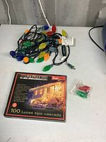 Photo showing large multicolored Christmas lights tangled on a table with a box of 'Everlife' 100 cascade lights and a small sealed pack of replacement bulbs.