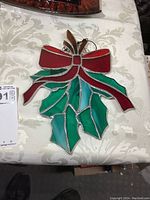 Full view of stained glass sun catcher showing red bow, green holly leaves, and brown accent leaves on a patterned white fabric background.