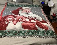 Photo of the full Christmas throw blanket laid flat showing the Santa Claus design holding toys and the holly border with fringed edges.