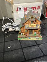 Front view of ceramic Christmas village cabin with power cord next to box labeled 'Gigama'