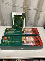 Three boxes of Holiday Collection outdoor illuminated garland, two 9 ft with 35 mini lights, one 18 ft, displayed on table with packaging visible showing product details.