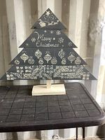 Front view of the solid wood Christmas tree decoration showing stencilled white designs and 'Merry Christmas' text on dark grey painted wood, supported by natural wood stand.