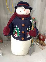 Full view of the large plush snowman wearing navy blue coat and maroon scarf, holding wood shovel decorated with greenery and berries