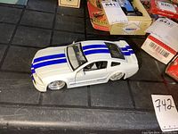 Front-left angled view of the white Mustang GT diecast model with blue racing stripes on a black grid surface.