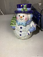 Front view of ceramic snowman cookie jar with purple top hat, blue bow tie, blue gloves holding candy cane and small Christmas tree decorations.