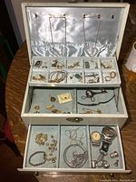 Open vintage jewelry box showing satin interior with compartments filled with assorted jewelry including necklaces, rings, pins, earrings and watches. Bottom and middle drawers visible with more jewelry items.