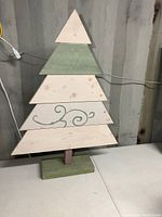 Front view of hand painted solid wood tree showing triangular layered panels in green and white with snowflake and swirl designs, standing on a rectangular base.