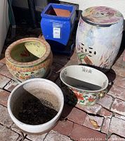 Five decorative planters grouped outdoors on brick surface showing variation in color and design.