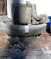 Front view of large oval cast cement water feature on pedestal with oval riser. Frog figure water spout pouring water. Small bucket of stones and small hollowed rock placed in front.