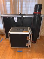 Four carrying cases grouped together: large black rectangular carrying case; glass display case with gold frame; black wheeled carrying case with telescoping handle; round black carrying tube with shoulder strap.