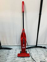 Front view of the red Simpli Stik bagless stick vacuum cleaner showing body, handle, base, and power cord on light carpet.