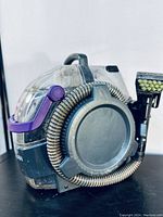 Front and side view of the BISSELL SpotClean Pet Pro showing the clear water tank, built-in hose, carry handle, and purple accent details on a black body.