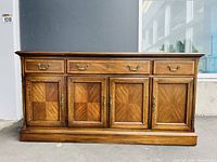Front view of solid wood sideboard showing four cabinet doors with decorative wood inlay and three drawers with brass handles.