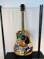 Front view of acoustic guitar covered in various stickers, showing natural wood grain and matte finish, set against a white background.