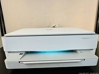 Front view of white HP ENVY 6024 wireless printer with blue light glowing inside output tray.