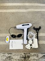 Top-down photo showing the Lescolton handheld laser device along with black protective goggles, power cable, instruction manual, and white laser attachment pieces arranged on a carpeted floor.