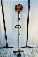 Front view of the weed wacker showing orange engine housing, gray shaft, and rusted metal blade with guard.