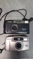 Both Canon Sure Shot ES Date (black) and Pentax Espio 738S (silver) cameras shown together on grey surface with wrist straps.
