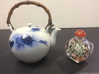 Front view of the round white ceramic teapot with blue decoration and bamboo handle next to a painted glass perfume bottle with orange stopper.