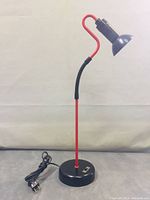 Full view of Kovacs post modern desk lamp showing black base, red rod, flexible neck, red curved segment and black lampshade, with power cord attached