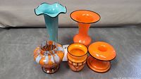 All five pieces of glassware displayed together on a gray surface, showing variety of sizes and shapes, predominantly orange pieces with one tall blue vase