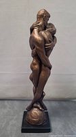 Side view showing the embracing couple with detailed anatomy, mounted on a round base on a black rectangular plinth.