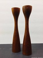 Full view of both wooden teak candle holders side by side on a dark surface, showing their tall, slim silhouette against a plain white background.