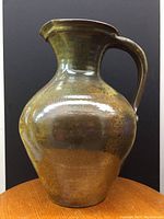 Full side profile of vintage large pottery jug showing its glossy brown and green earthy glaze, curved handle, and broad round body with a flaring neck and spout.