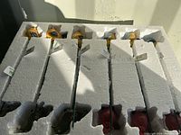 View of 12 Evergreen plant picks secured in white foam packaging showing the tops of colorful bird decorations attached to stakes.