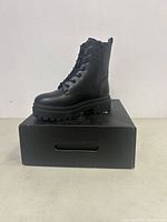 Side view of a black combat boot showing lace-up front, side zipper, heel pull tab, and chunky sole on a black box with Fashion Nova branding.